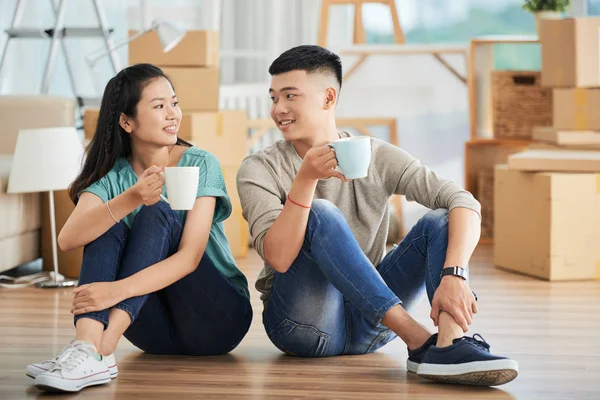 愉快的年轻亚洲夫妇坐在一起 在地板上喝着一杯热饮 面面相觑 — 图库照片