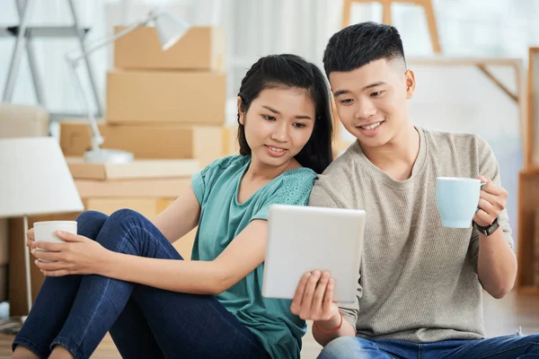 幸せな若いアジア男と女マグカップで飲み物の新しいアパートの床に座って タブレットを見て — ストック写真
