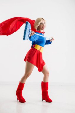 Genç güzel sarışın süpürge tutarak ve neşeyle beyaz arka planı gülümseyen superwoman kostüm tam uzunlukta portresi