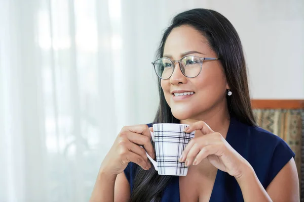 飲み物のカップを保持しているとしんみりと離れた事務所で椅子に座ってお探しのメガネで大人のタイの女性 — ストック写真
