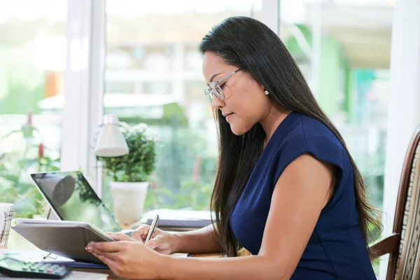 亚洲成年妇女在纸上写字 看平板电脑在灯房的桌子上工作的侧视图 — 图库照片