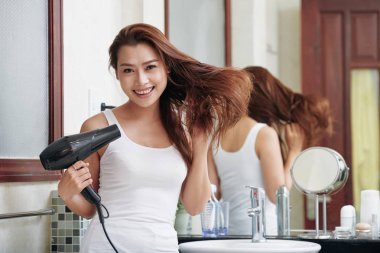 Young charming Asian woman drying hair and smiling at camera standing in bathroom
