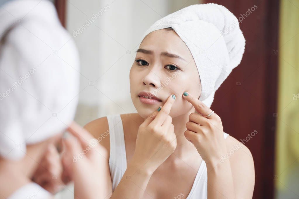 Young Asian woman in towel standing in front of mirror and popping pimples looking concerned