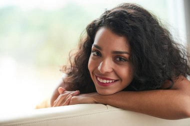 Onun kanepenin arkasına yaslanmış ve kameraya bakarak çekici gülümseyen genç kadın