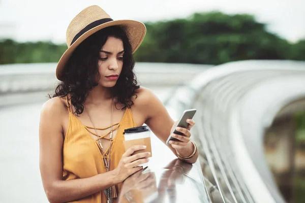 Pretty frowning young Hispanic woman in straw hat standing outdoors with cup of coffee and checking phone