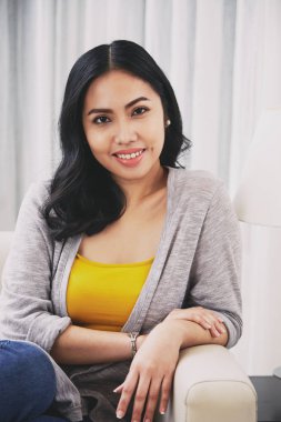 Gorgeous Filipino female smiling and looking at camera while relaxing on comfortable sofa in cozy living room