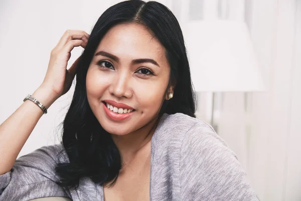 아늑한 거실에 앉아있는 카메라를보고 머리를 만지는 매력적인 미소와 아름다운 필리핀 — 스톡 사진
