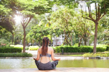 Arkadan görünüşü anonim esmer Teras park gölet güneş ışığı altında oturan ve meditasyon spor giyim