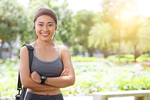 身穿运动服的年轻亚洲女人提着包 戴着耳机 双手交叉地对着相机微笑 — 图库照片