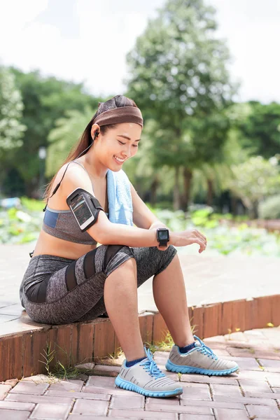 Young Asian woman in sportswear and with armband and smart watch sitting in summer park and smiling