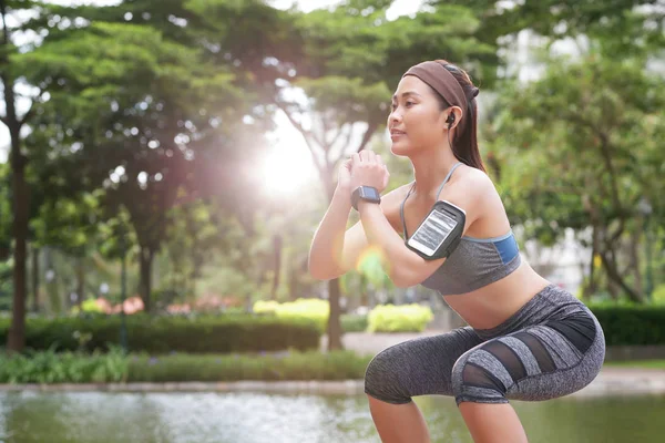 自信的年轻亚洲女运动员在袖标和健身跟踪器做蹲和锻炼在阳光下 — 图库照片