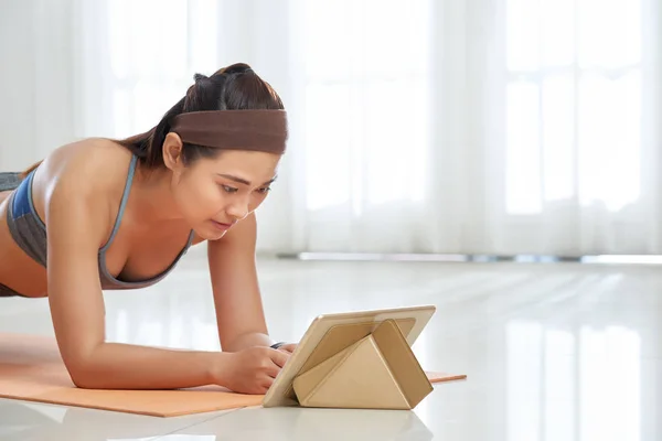 年轻的亚洲女运动员在垫子上做木板运动 在家看平板电脑 — 图库照片