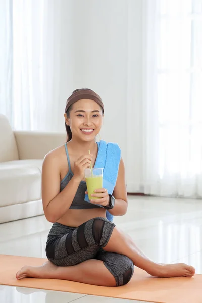 自宅のマットの上に座って陽気なアジア スポーツウーマン緑のスムージーのグラスを持って カメラで笑顔 — ストック写真