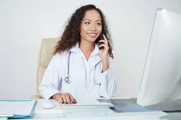 微笑的年轻专业女医生坐在她的工作场所 在电脑打字 而用手机交谈 — 图库照片