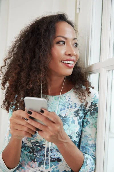 Kıvırcık Saçlı Güzel Kadın Akıllı Telefonundan Müzik Dinleyip Pencereden Bakıyor — Stok fotoğraf