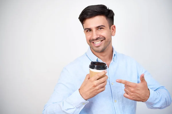 コーヒーの使い捨てカップと立っている白い背景に分離されてそれを指して幸せの若い男の肖像 — ストック写真