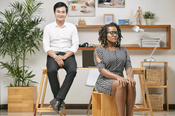 彼女の後ろに彼女の若いアジアの従業員とオフィスで座っている眼鏡で自信を持ってアフリカの女性指導者 — ストック写真