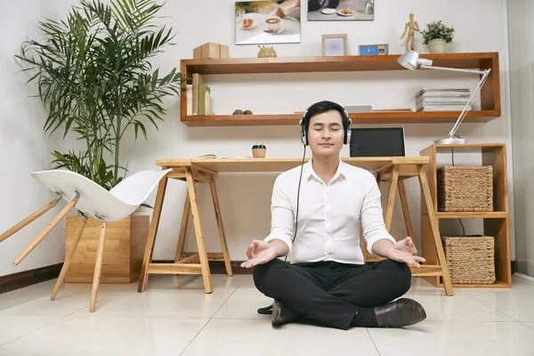 若いアジア系のビジネスマンをヘッドセットを着てオフィスで彼の職場の近くの床の上に座って音楽を聴くと 休憩中に瞑想 — ストック写真
