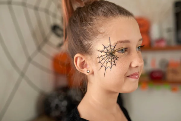 Half-turned portrait of little pretty Caucasian girl with makeup and painted   Halloween spider web on her face looking away and smiling