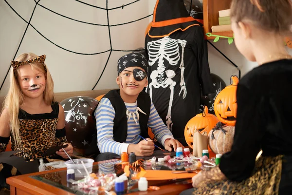 海賊衣装とテーブルに座って ハロウィーンの飾りを作りながらカメラ目線の猫衣装で彼の女友達で白人少年 — ストック写真
