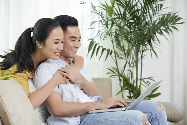 年轻的亚洲夫妇坐在家里的沙发上冲浪笔记本电脑的侧视图 — 图库照片
