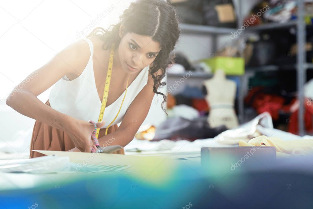 Young fashion designer using scissors to cut piece of cloth on table while working in tailor workshop