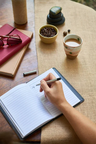 她闭上了头 拿着笔 在日记里写着新的想法 坐在桌旁 拿着一碗咖啡豆和一杯咖啡 — 图库照片
