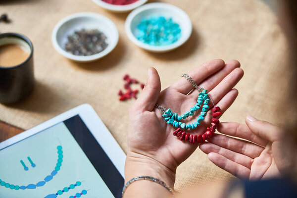 Close-up of woman weaving necklace from natural stone beads using sample on tablet pc, she making the same necklace from different stones