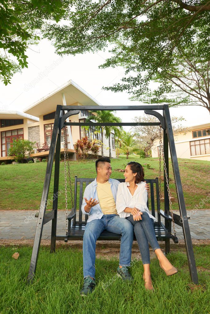 Happy Asian couple sitting on swing talking and smiling with their beautiful house in the background