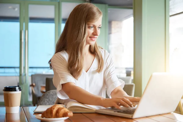 美丽的女人打字在笔记本电脑键盘上的咖啡馆桌子与咖啡和羊角面包 — 图库照片