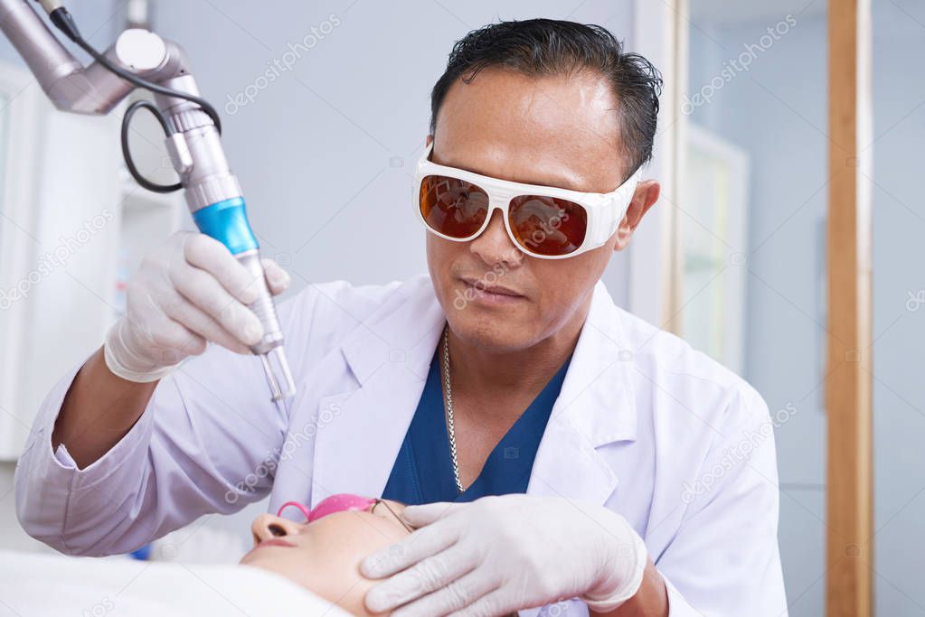 Male specialist in protective glasses lifting laser rejuvenation device under female face at beauty salon