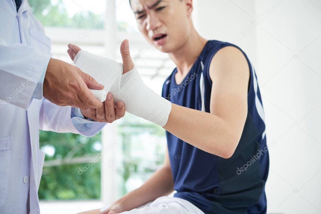medical practitioner applying clean bandage on injured arm of young Asian guy