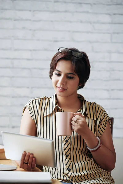 Smiling young woman drinking coffee and reading data on tablet screen during short break