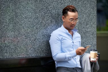 Duvara yaslanmış, kahve içme ve telefon metin okuma yakışıklı orta yaşlı işadamı
