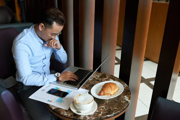 Pensive Vietnamese businessman having brunch in cafe and working on laptop