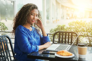 Gülümseyen ve güneşli günde modern dizüstü yakınındaki kafede masada otururken kameraya bakarak güzel Endonezya Bayan yan görünüm