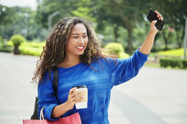 漂亮的印尼女性与一杯热饮微笑和挥舞着手与智能手机 而站在模糊的背景街道在购物 — 图库照片