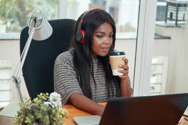 漂亮的年轻黑人妇女在耳机喝咖啡和在办公室的笔记本电脑工作 — 图库照片