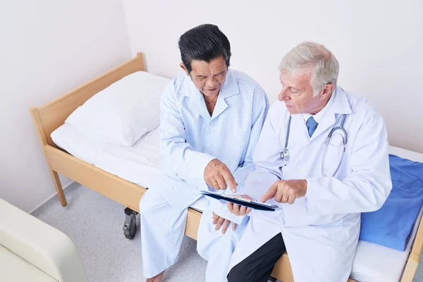上記の高齢医師と病棟でベッドの上に座っている間現代タブレットを指してのアジアの患者のショット — ストック写真