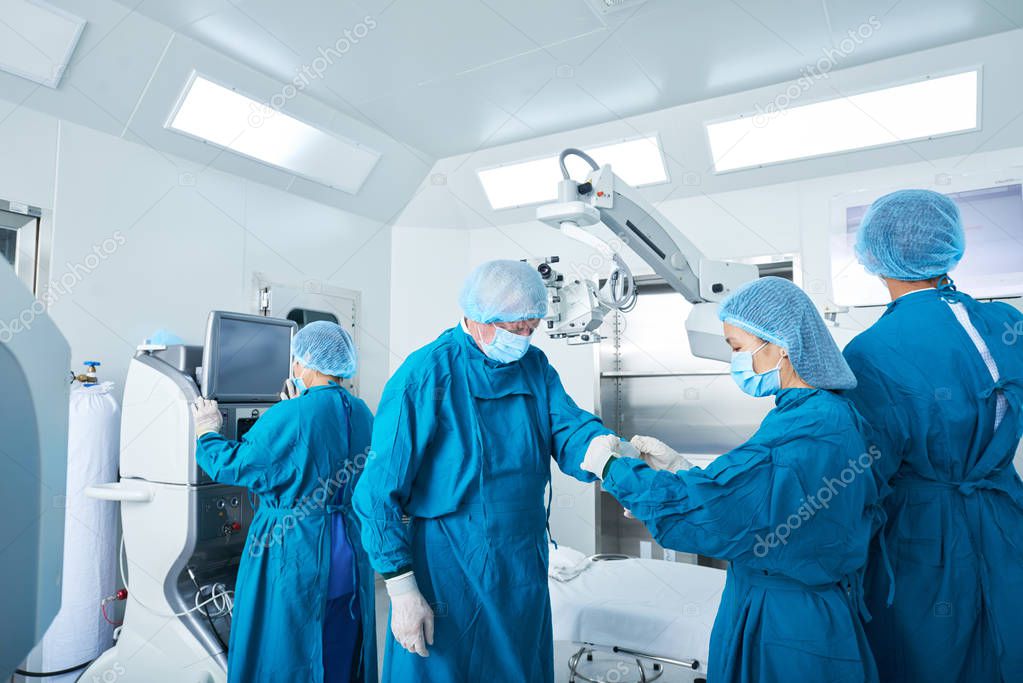 Young surgical assistant helping experienced surgeon to put on latex gloves before performing surgery in operating theater