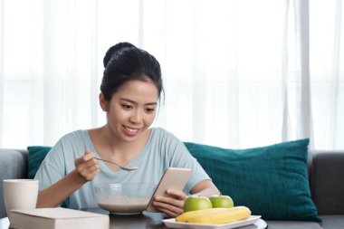 Ne zaman kahvaltı yemek Genç Asyalı kadın kontrol sosyal medya Smartphone cep telefonu ile gülümseyen