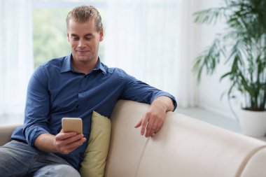 Kanepede oturan ve onun smartphone içinde metin mesajları okuma gülümseyen yakışıklı genç adam