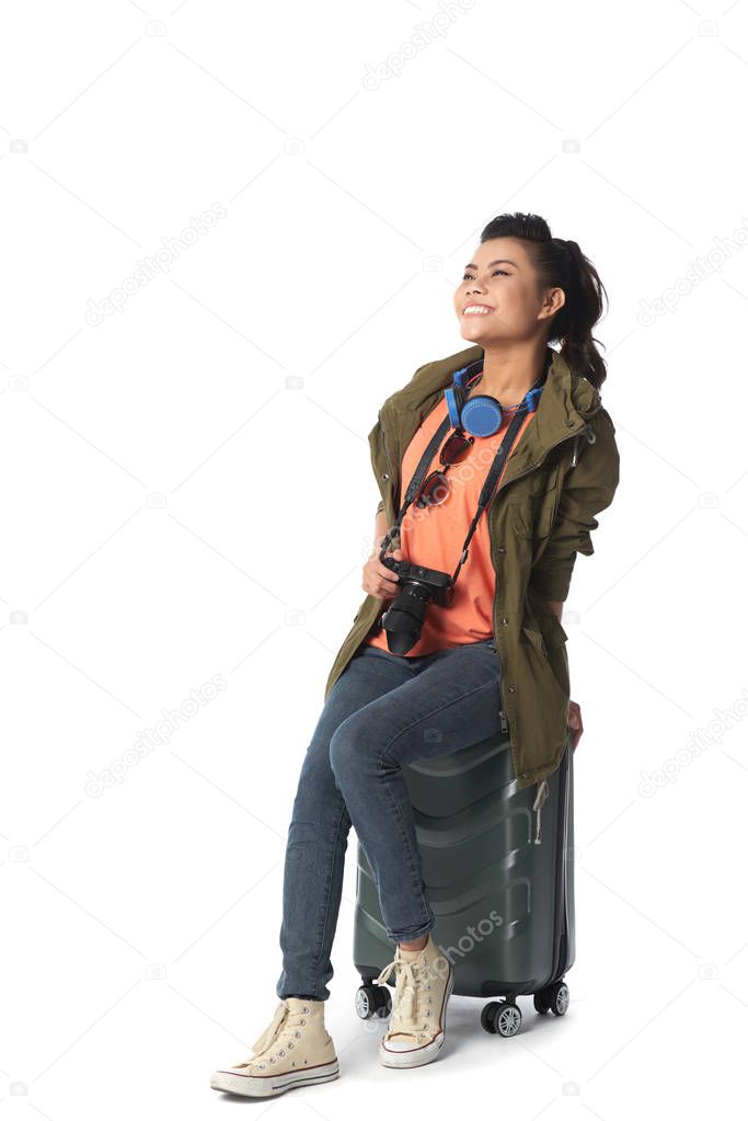 Excited happy Vietnamese young woman sitting on suitcase, isolated on white