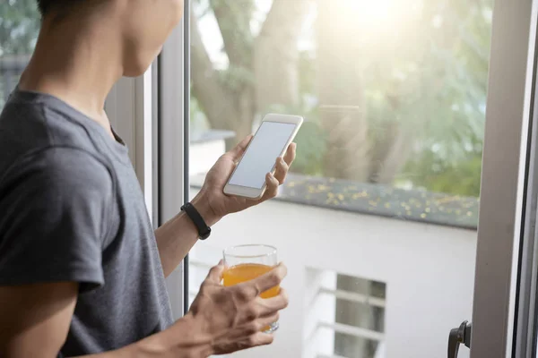 Young man drinking of juice and checking weather forecast on smartphone when standing at window