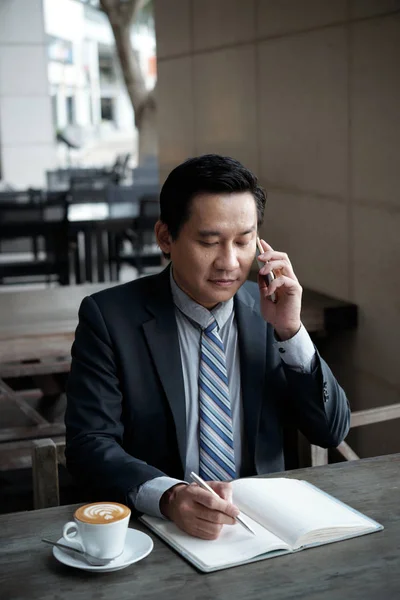 Azjatycki Biznes Executive Pytając Telefon Robienia Notatek Podczas Picia Kawy — Zdjęcie stockowe