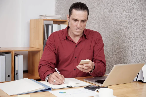 Busy office worker looking at smartphone while making notes in his notepad at office