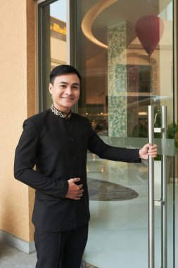 Asian man in elegant black suit opening glass doorway of hotel greeting  clipart