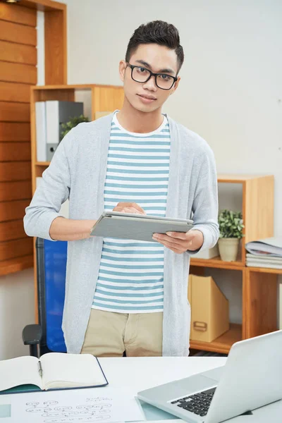 穿着眼镜的帅气亚洲人 用平板电脑站在桌旁 在办公室看着相机 — 图库照片