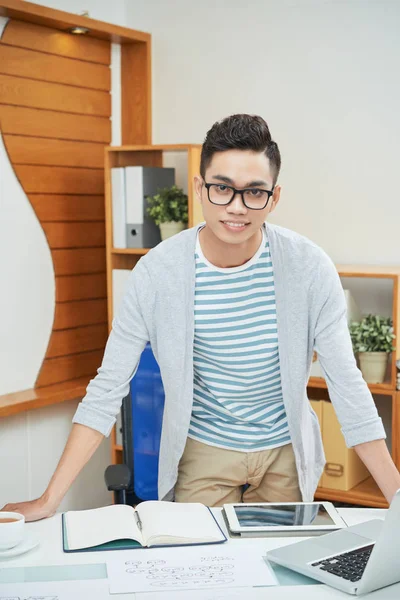 Confident Asian man in glasses standing at table in contemporary office leaning on table and looking at camera
