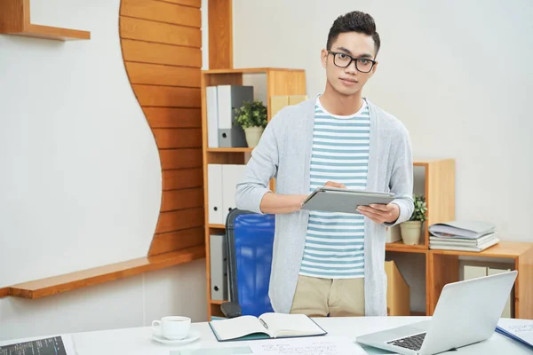 年轻的休闲亚洲男子戴眼镜站在平板电脑在现代办公室看相机 — 图库照片